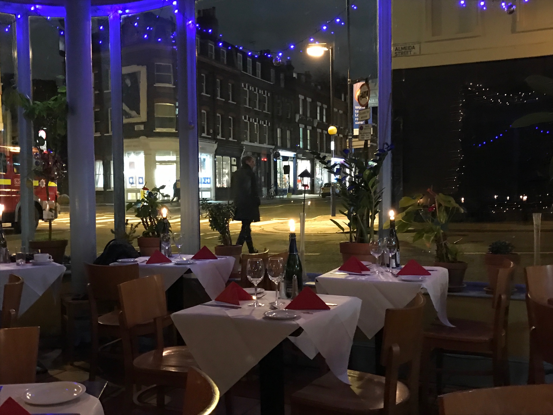Restaurant tables with red napkins beside a window over an Islington street at night